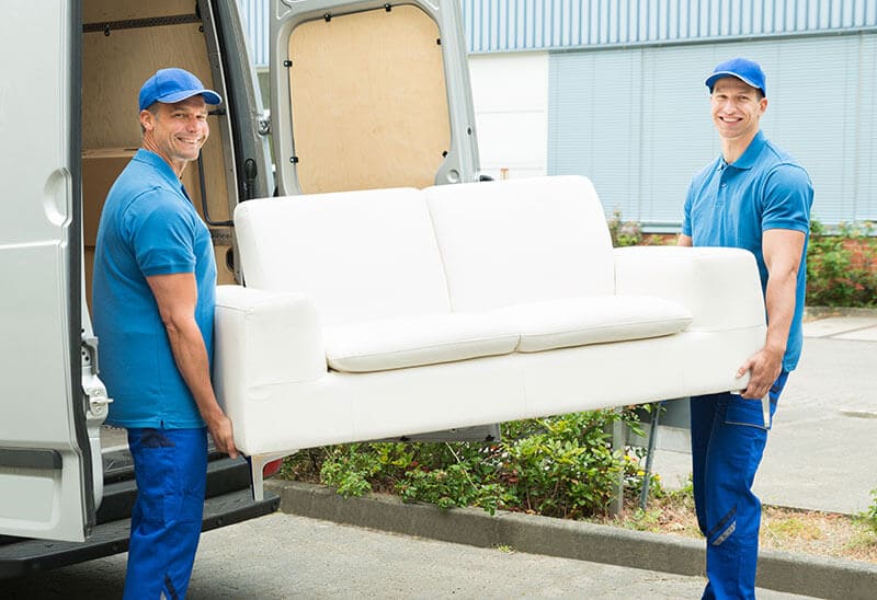 The image displays the front section of a white Hire Man and Van removal van parked on a residential driveway in Hockley, with a driveway surface of grey concrete visible in the foreground. The van's side panel, featuring a smooth, clean finish with faint reflections from overcast sky, occupies the left side of the image. Part of the van’s front wheel and a portion of the bumper are visible, with the bumper painted in a matching white. In the background, a portion of a brick-built house with a small front garden and a wooden fence can be seen, suggesting a typical suburban setting. The scene is illuminated by diffuse daylight, creating soft shadows and neutral tones throughout. The van appears to be prepared for rubbish removal or moving tasks, consistent with local waste management services in the SS1 postcode area. The overall setting emphasizes a practical, residential environment where Hire Man and Van provides rubbish collection and removal services to households in Hockley. No waste or other objects are visible in the immediate vicinity of the vehicle in this shot, focusing solely on the vehicle's appearance and context within a local residential street.